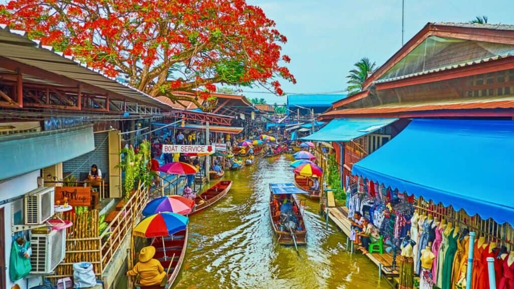 The colorful umbrellas, canopies and boats of Damnoen Saduak floating market with narrow khlong and blooming flame tree over the roofs - Stock